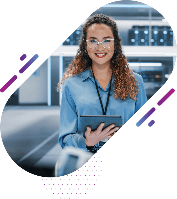 A smiling woman with curly hair and glasses, wearing a blue shirt and holding a tablet, standing in a modern data center environment with servers in the background.